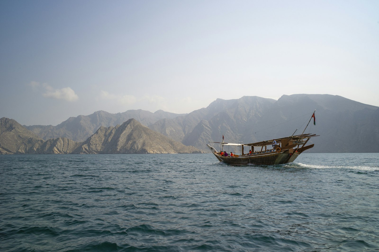 Traditional wooden Omani dhow flying the national flag, sailing across the calm waters of the Musandam fjords beneath rugged mountains